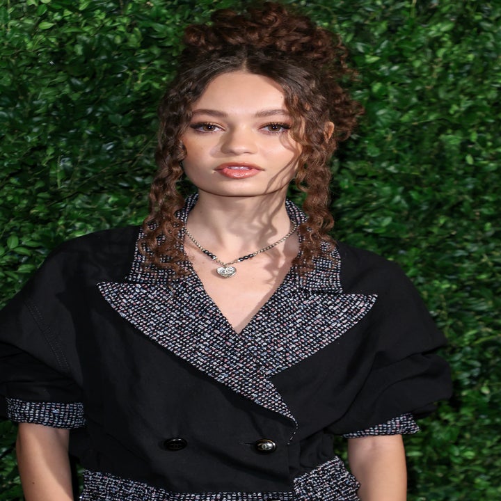 Nico Parker in a stylish, textured two-piece outfit with curly hair stands in front of a green foliage backdrop