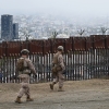 In this photo, two members of the U.S. Marine Corps patrol on foot near a U.S.-Mexico border fence. They are wearing uniforms, boots and helmets. Behind the fence, buildings in Tijuana, Mexico, rise up in the background.