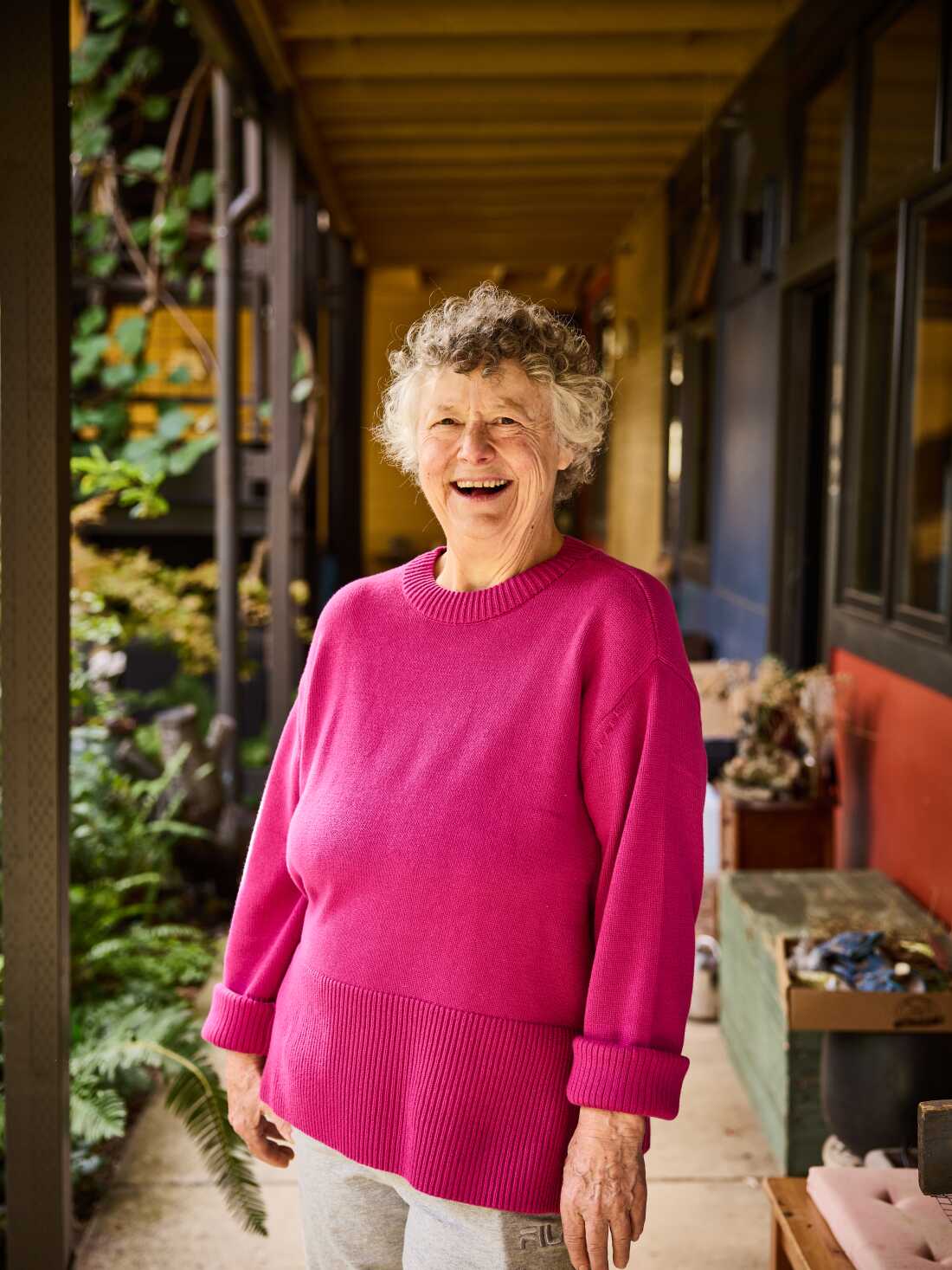 Deana Camp, 73, has curly gray hair and is wearing a bright pink sweater. She is smiling broadly.