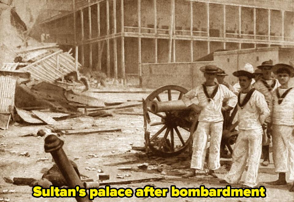 Historic photo of sailors in uniform standing near a cannon amid debris, possibly from a past conflict scene
