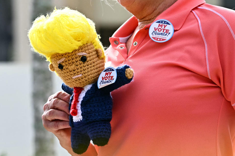 A person in a collared shirt holds a crochet doll with yellow hair, wearing a suit. Both wear "My Vote Counts!" buttons