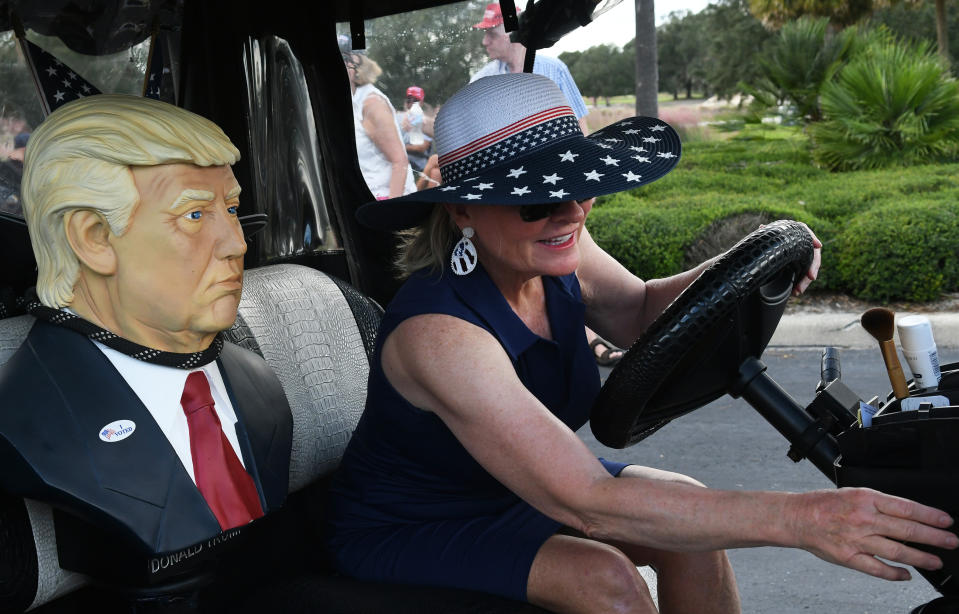 A woman in a large sunhat drives a golf cart with a mannequin bust of Donald Trump in a suit and red tie seated beside her