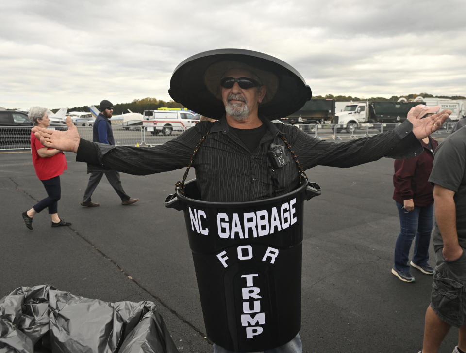 Man wearing a black garbage can costume that reads "NC Garbage for Trump," with arms outstretched, at an outdoor event