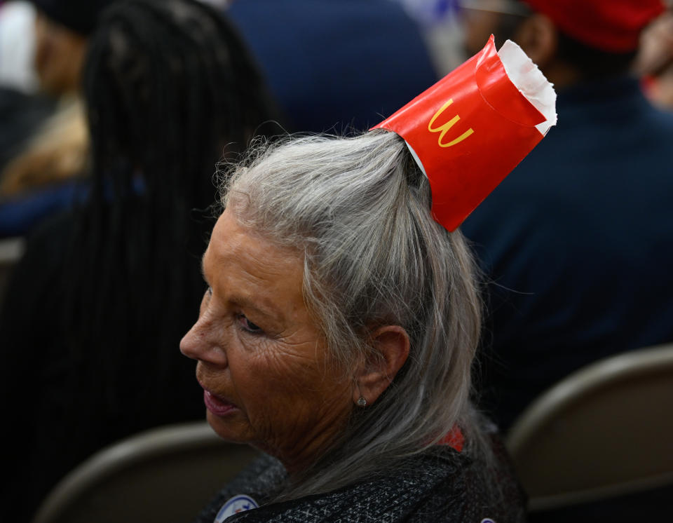 A person with silver hair wearing a McDonald's fries container as a hat, seated among a crowd