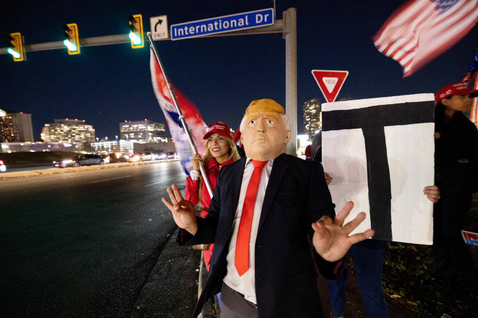 A person in a Trump mask gestures with their hands on a street at night. Others are nearby with signs, under a street sign
