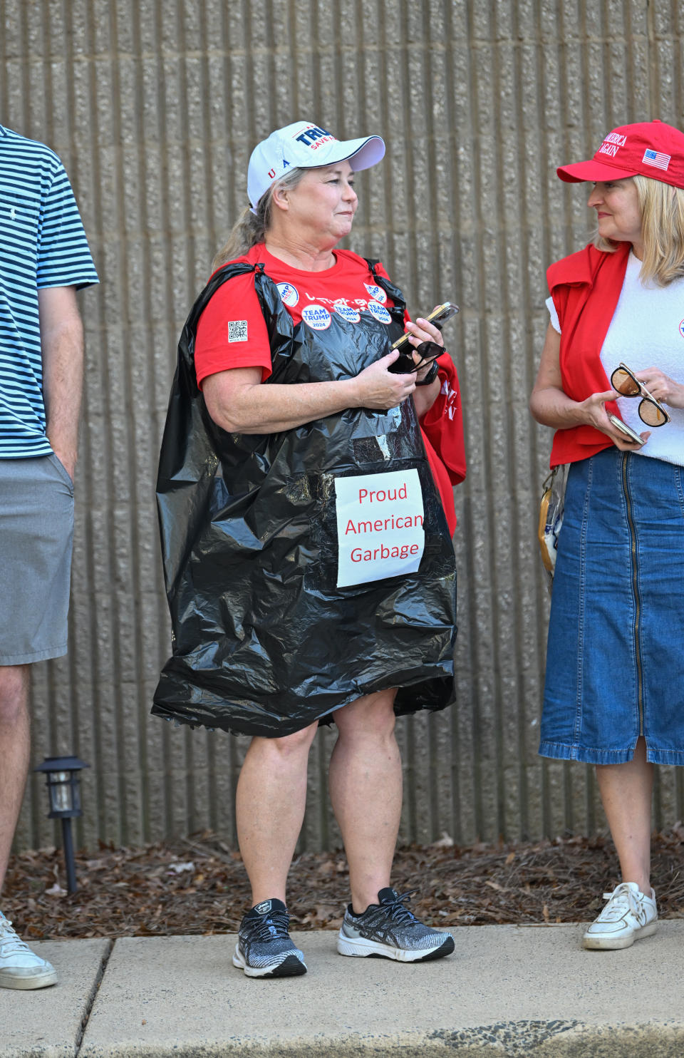 A person wearing a black trash bag with a "Proud American Garbage" sign at an outdoor event. They're wearing a hat and a red shirt