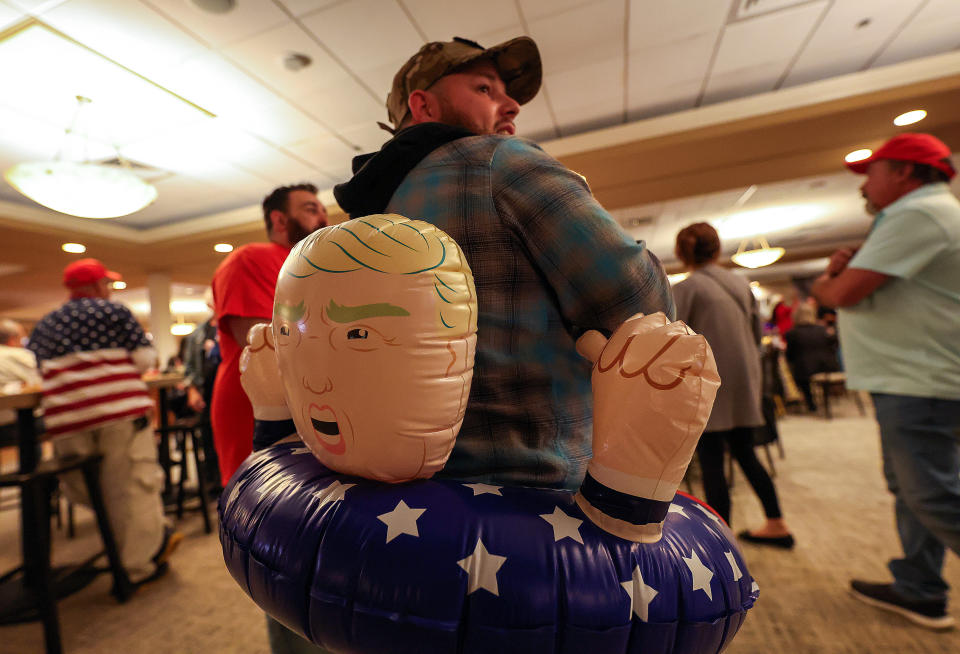 Person in a plaid shirt wearing a Trump inflatable pool float around their waist in a crowded indoor setting