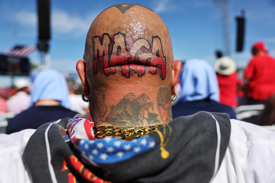 Person with "MAGA" head tattoo at an outdoor event, wearing a necklace, facing the stage