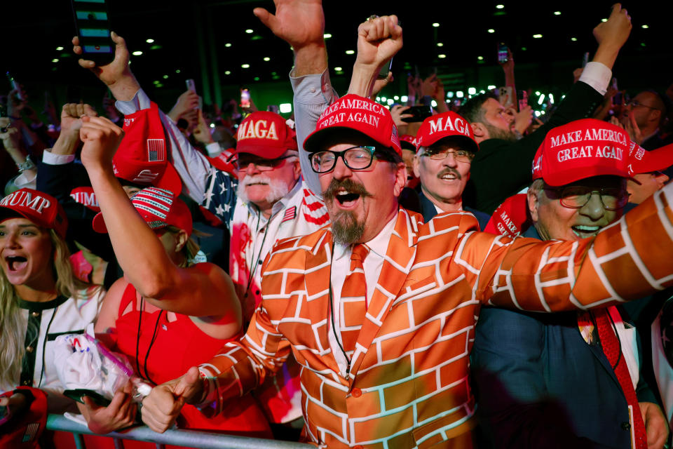 People in crowd wearing "Make America Great Again" hats, with one person in a brick-pattern suit, cheering enthusiastically