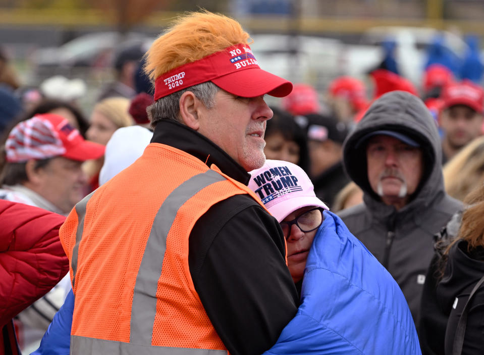 A man in a "Make America Great Again" visor hugs a woman wearing a "Women for Trump" hat in a crowded outdoor event