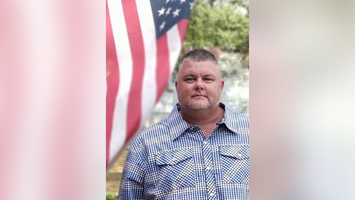 The late George Garner II, former mayor of McColl, South Carolina, is seen wearing a blue and white checked shirt while standing in front of an American flag outdoors.